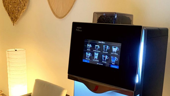Automat kawowy w recepcji Strefy Saun. Dotykowy wyświetlacz z dostępnymi do wyboru kawami. Po jego lewej stronie ratanowy fotel, a nad nim wiklinowe wachlarze dekoracyjne. 