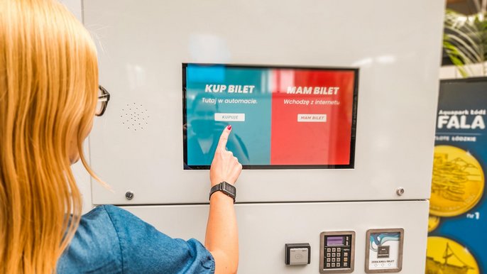 Blondynka stojąca tyłem do obiektywu stoi przed biletomatem - wyciągniętym palcem wskazuje na opcję "KUPUJĘ". 