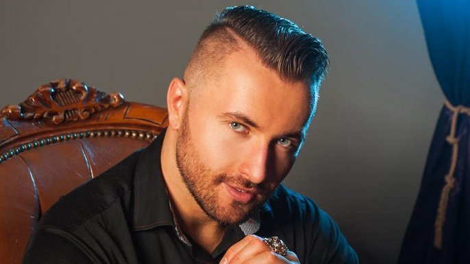 Saunamistrz Damian Białas - młody mężczyzna siedzi na stylizowanym, skórzanym fotelu. Spogląda w obiektyw, opiera brodę na splecionych dłoniach. Ma na sobie czarną koszulę. 