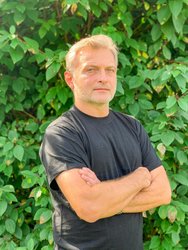 Mężczyzna w średnim wieku ubrany w czarną koszulkę, ręce ma splecione na klatce piersiowej. Jasne włosy i jasne oczy, lekki uśmiech.