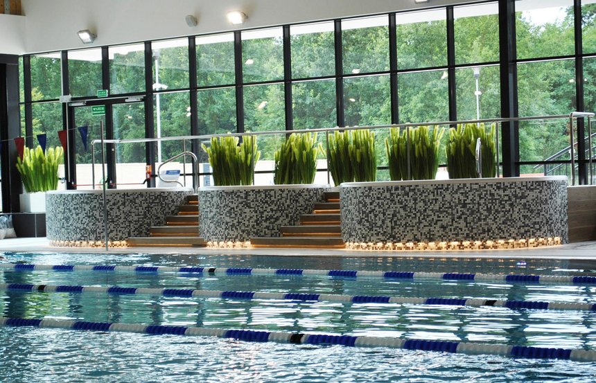 Trzy wanny jacuzzi obłożone szarą mozaiką ceramiczną umiejscowione wzdłuż przeszklonej ściany hali. Przed wannami widać część basenu sportowego, za nim donice z zieloną roślinnością.