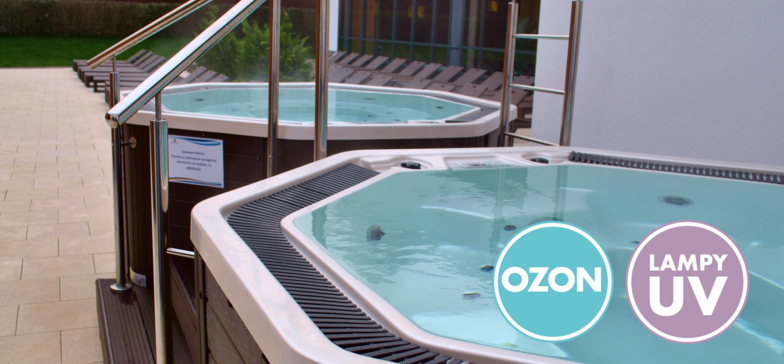 Dwa zewnętrzne jacuzzi w Strefie Saun obudowane ciemnobrązową okładziną imitującą drewno. W prawym dolnym rogu znaki informacyjne: OZON i LAMPY UV.
