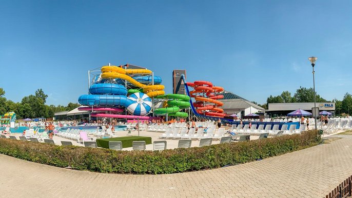 Panorama terenu zewnętrznego w słoneczny dzień, na pierwszym planie niski zielony żywopłot, w centrum kolorowe zjeżdżalnie, po lewo baseny sportowy i rekreacyjny, a po prawo basen wypływowy z halą z logotypem FALA. 