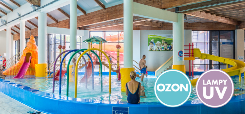 Wodny Plac Zabaw z Muszlą. Przy brzegu mama obserwująca zabawy dzieci. W tle pomarańczowa muszla z małą zjeżdżalnią. W prawym dolnym rogu znaki informacyjne: OZON i LAMPY UV.
