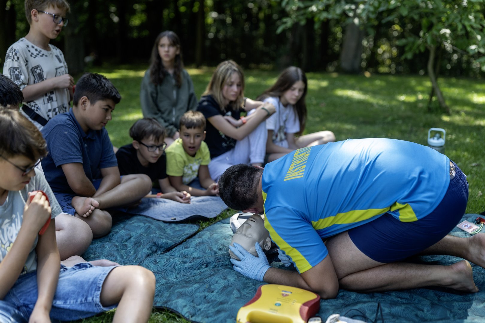 Grupka dzieci ćwicząca pierwszą pomoc. Na pierwszym planie jest instruktor. 