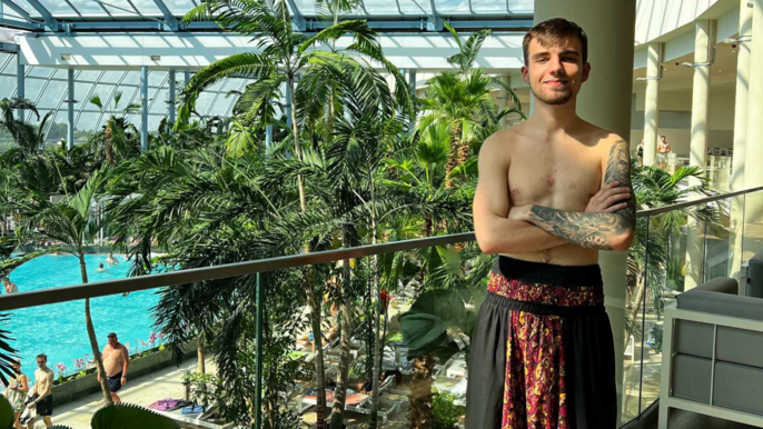 Uśmiechnięty brunet z delikatnym zarostem. Pozuje w budynku, na tle palm. W oddali widać fragment basenu. Mężczyzna jest bez koszulki. Jest ubrany we wzorzyste luźne spodnie haremowe. 