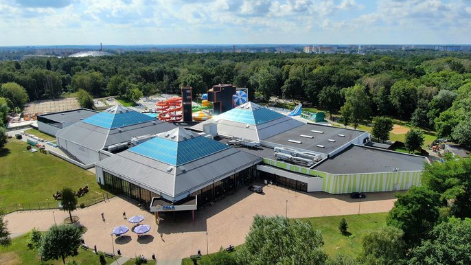 Panorama Aquaparku widoczna od strony wejścia głównego. Trzy przeszklone piramidy Aquparku otoczone zielonym parkiem. 