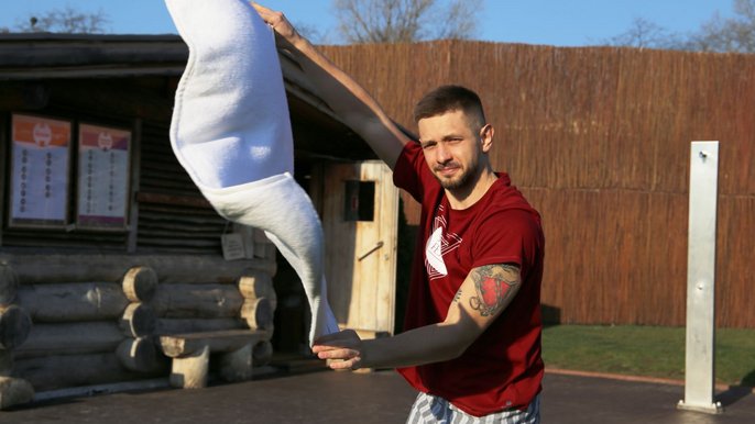 Saunamistrz Michał Blejder brunet z zarostem ubrany w bordowy t-shirt i ręcznik w paski. Pozuje na tle wiklinowego płotu. Macha białym ręcznikiem. W tle zewnętrzna sauna będąca drewnianym budynkiem. 