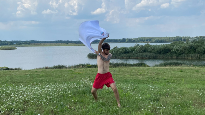 Mężczyzna bez koszulki, w czerwonych spodenkach przed kolana. Pozujący na terenie łąki. W tle widoczne jezioro. W górze trzyma rozłożony biały ręcznik. 