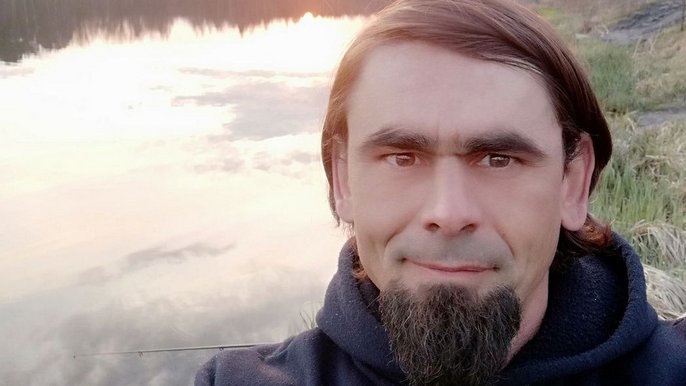 Saunamistrz Michał Rzewuski - brunet z ciemną brodą poluje na tle jeziora oświetlonego wschodem słońca. 