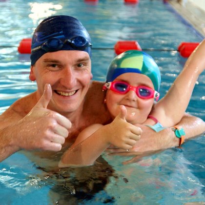 Zbliżenie na instruktora obejmującego kilkuletnią dziewczynę w okularkach i czepku pływackim. Oboje szeroko uśmiechnięci pokazują uniesione do góry kciuki.