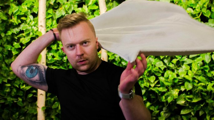 Saunamistrz Sebastian Dziurski. Blondyn w czarnej koszulce unosi za plecami rozłożony ręcznik biorąc zamach. Na lewym ramieniu ma tatuaż kolorowego oka. Pozuje na tle ściany zielonej roślinności. 