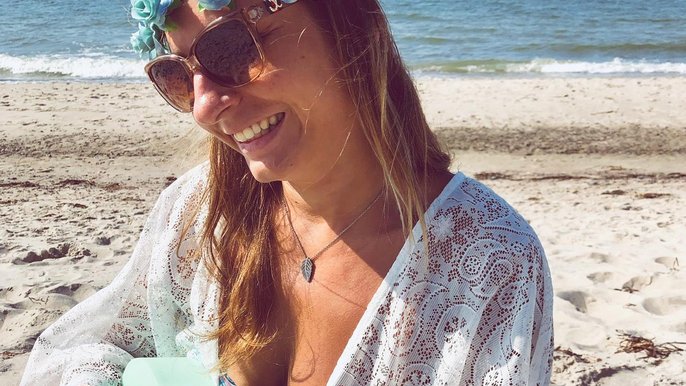 Saunamistrzyni Mariola Żurek pozująca na plaży na tle morza. Ma na sobie wianek z niebieskich kwiatów i zwiewną biała koszulę w drobny wzór. W ręku trzyma miętową małą gitarę. 