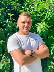 Młody uśmiechnięty mężczyzna o silnej budowie ciała. Ma na sobie białą koszulkę z napisem Aquapark FALA. Blondyn o jasnych włosach ma ręce zaplecione na klatce piersiowej, na jednym nadgarstku nosi zegarek.
