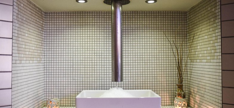 Biała umywalka umieszczona pod aluminiową rurą, którą opada lód z łuskarki. Wokół jasne płytki.