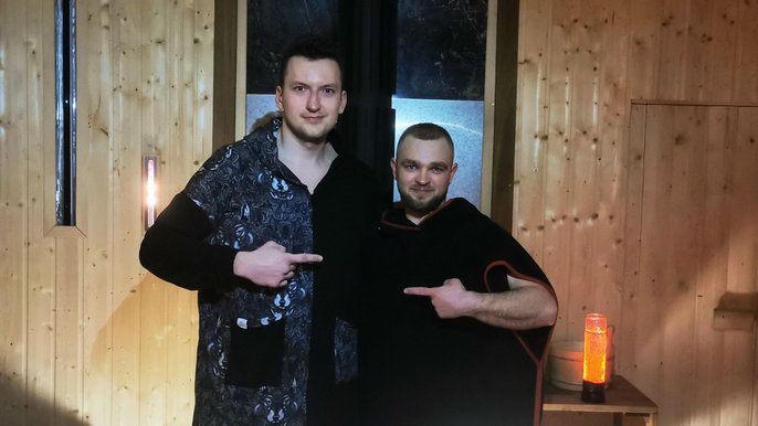 Saunamistrzowie Dominik Grala (po lewej) i Artur Podwójci (po prawej). Mężczyźni pozują w saunie. Ubrani w peleryny saunowe. Stoją obok siebie i wskazują na siebie. 