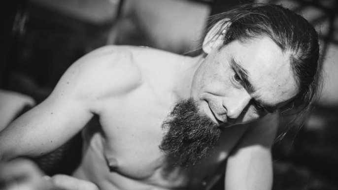 Czarno-białe zdjęcie. Saunamistrz Michał Rzewuski, brunet z ciemną brodą, bez koszulki. Mężczyzna pozuje patrząc na niewidoczny na zdjęciu obiekt. 