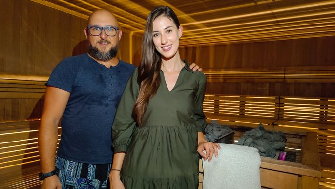 Saunamistrzowie Marek Olszewski i Kasia Kluba. Uśmiechnięty duet pozujący na tle sauny fińskiej. Kobieta ubrana w oliwkową tunikę. Mężczyzna w okularach, ubrany w granatowy tshirt i granatowe wzorzyste spodnie. 