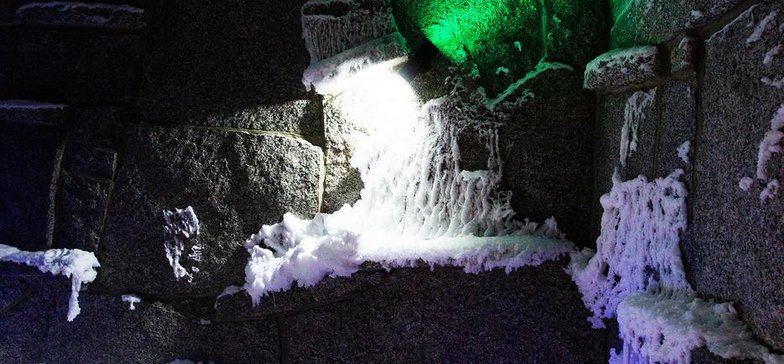 Wnętrze groty śnieżnej - ciemne kamienie na ścianach, warstwa śniegu na podłodze, wiszące stalaktyty z sufitu pokryte są śniegiem. Resztki śniegu znajdują się również na ścianach. Oświetlenie w kolorze zielonym i niebieskim.
