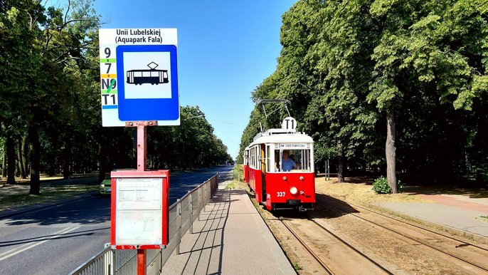 Przystanek tramwajowy przy al. Unii Lubelskiej. Po lewej asfaltowa droga, wzdłuż niej liczne drzewa. Na przystanek podjeżdża zabytkowy czerwony tramwaj z białymi elementami dachu i obramowania okien. 