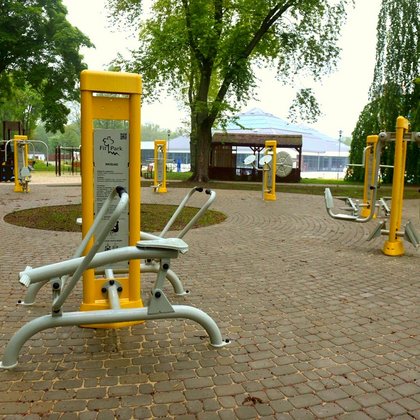 Siłownia zewnętrzna usytuowana w części parkowego Aquaparku FALA. Na szarej kostce brukowej stoją żółto-szare sprzęty. Na pierwszym planie wioślarz, w tle plac zabaw dla dzieci i jedna z hal-piramid Aquaparku.