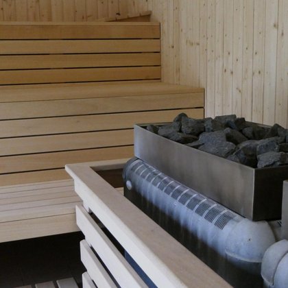 Wnętrze małej sauny zewnętrznej, zbliżenie na podwójny prostokątny piec z ułożonymi ciemnymi kamieniami. W tle trzy rzędy drewnianych ław.
