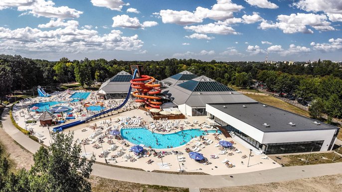 Panorama terenu zewnętrznego w słoneczny dzień uchwycona z perspektywy drona. Na pierwszym planie basen wypływowy i hala z basenem sportowym, dalej wieża zjeżdżalni oraz baseny sportowy i rekreacyjny. Świetnie widać hale w kształcie piramid. 