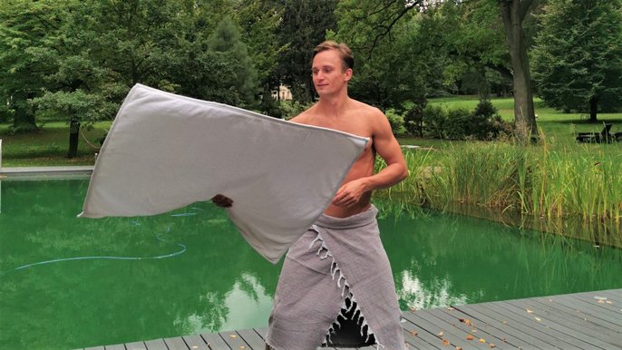 Saunamistrz Bartosz Klag. Mężczyzna o jasnych włosach, bez koszulki, w szarym ręczniku. Macha białym ręcznikiem, w parku, na tle drzew i zbiornika wodnego. 