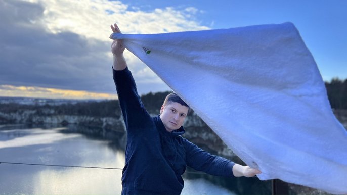 Saunamistrz Sebastian Domański, brunet o ciemnych oczach ubrany w czarną bluzę. Pozuje na tle jeziora i lasu. Na zdjęciu uchwycone jest machanie białym ręcznikiem przez Sebastiana. 