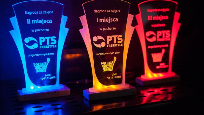 Trzy szklane statuetki oświetlone kolejno od lewej: niebieskim, pomarańczowym i czerwonym strumieniem światła. Statuetki są nagrodą w turnieju PTS Freestyle, mają na sobie logo turnieju i Polskiego Towarzystwa Saunowego. 