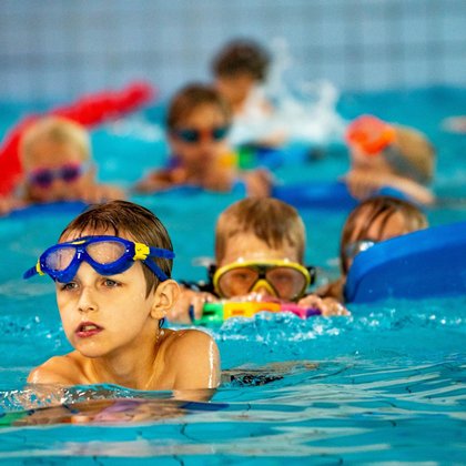 Zbliżenie na dzieci na jednym z torów w basenie sportowym. Na pierwszym planie chłopiec z okularkami pływackimi przesuniętymi na czoło. W tle kilkoro innych dzieci w okularkach i z deskami pływackimi.