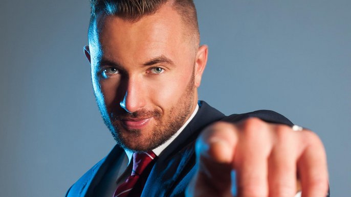 Młody mężczyzna w ciemnym garniturze i czerwonym krawacie patrzy wprost w obiektyw wskazując w jego stronę palcem. Włosy w kolorze ciemny blond ma krótko przystrzyżone, na twarzy kilkudniowy zarost. 