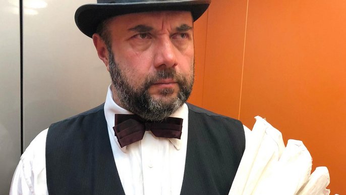 Saunamistrz Wojciech Draga w meloniku, czarnej kamizelce i muszce oraz białej koszuli. Mężczyzna w średnim wieku z krótką brodą w rękach trzyma biały wachlarz saunowy. 