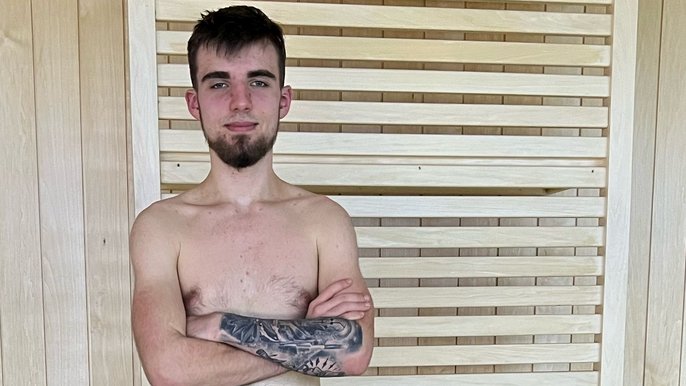 Sanamistrz Arkadiusz Dan, brunet z ciemną krótką brodą. Pozuje bez koszulki na tle drabinek sportowych i ściany z jasnego drewna. Na prawej ręce, z perspektywy zdjęcia, ma tatuaż. 