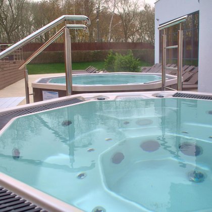 Dwie wanny jacuzzi obudowane ciemnym drewnem, zbliżenie na jedną z nich, widać parę unoszącą się nad wodą, pomiędzy nimi podest z aluminiową poręczą. W tle plaża terenu zewnętrznego Strefy Saun.