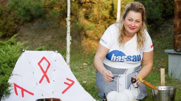 Saunamistrzyni Mirela Kazuś-Barańska. Blondynka pozuje w leśnym krajobrazie. Klęczy przy trzech śnieżnych kulach, które zakrapla olejkiem eterycznym. Obok wachlarz saunowy. 
