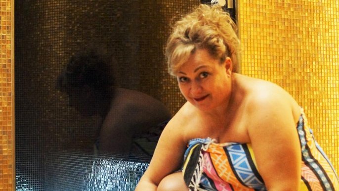 Blondynka w średnim wieku w kolorowym hammamie klęczy nad ceramicznym talerzem, na którym umieszczona jest lodowa kula. Wokół niej rozłożone są fiolki z olejkami aromaterapeutycznymi i wiadro i łyżka służące do polewania wodą pieców sauny. 