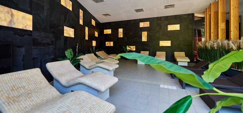 Wnętrze tepidarium. Beżowe, ceramiczne leżaki podgrzewane otoczone są ścianami wyłożonymi ciemnymi płytkami imitującymi kamień. Gdzieniegdzie na ścianie jasne panele świetlne.