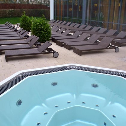 Teren zewnętrzny Strefy Saun widziany z perspektywy jacuzzi. Na pierwszym planie lustro wody jacuzzi, w tle plaża zewnętrzna z brązowymi leżakami, a dalej przeszklona ściana Strefy Saun.