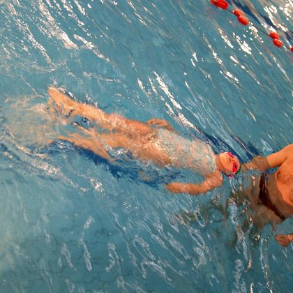 Instruktor stoi w basenie sportowym, przed sobą ma kilkuletnią dziewczynkę w błękitnym stroju kąpielowym, unoszącą się na wodzie na plecach.