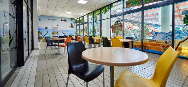 Bar Mokry przy basenie z falą - na pierwszym planie stoliki otoczone kolorowymi krzesłami. W tle fototapeta oraz przeszklona ściana ozdobione motywami pirackimi. Zza szyby widać most linowy i basen z falą.