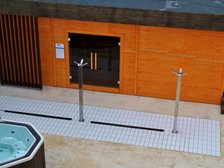 Duża sauna zewnętrzna z zewnątrz. Zabudowana drewnem w kolorze pomarańczowym, z brązowymi elementami dekoracyjnymi. Przed wejściem do sauny trzy poczwórne prysznice zewnętrzne i jacuzzi.