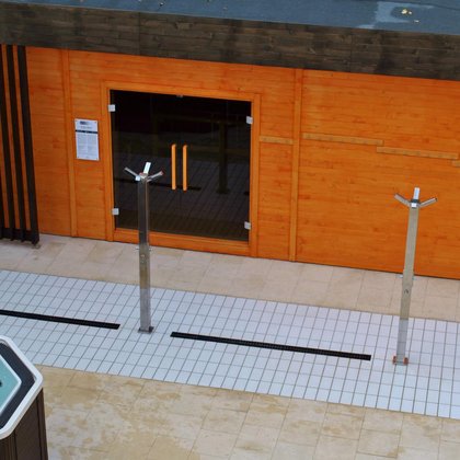 Duża sauna zewnętrzna z zewnątrz. Zabudowana drewnem w kolorze pomarańczowym, z brązowymi elementami dekoracyjnymi. Przed wejściem do sauny trzy poczwórne prysznice zewnętrzne i jacuzzi.