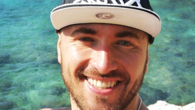 Saunamistrz Damian Białas. Uśmiechnięty blondyn z lekkim zarostem ma na sobie granatową czapkę z daszkiem, w tle lazurowa woda. 
