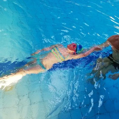 Instruktor stoi w basenie sportowym, przed sobą ma kilkuletnią dziewczynkę w dwuczęściowym stroju kąpielowym, unoszącą się na wodzie na plecach.
