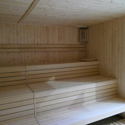 Wnętrze małej sauny zewnętrznej. Po prawej stronie przeszklone drzwi, a naprzeciw trzy rzędy drewnianych siedzisk. Cała sauna obudowana drewnem.