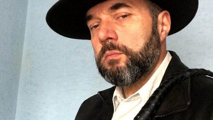 Saunamistrz Wojciech Draga - mężczyzna w średnim wieku z ciemną brodą ma na sobie strój inspirowany Indiana Jonsem: ciemny kapelusz i skórzaną kurtkę, a w dłoni bicz. 