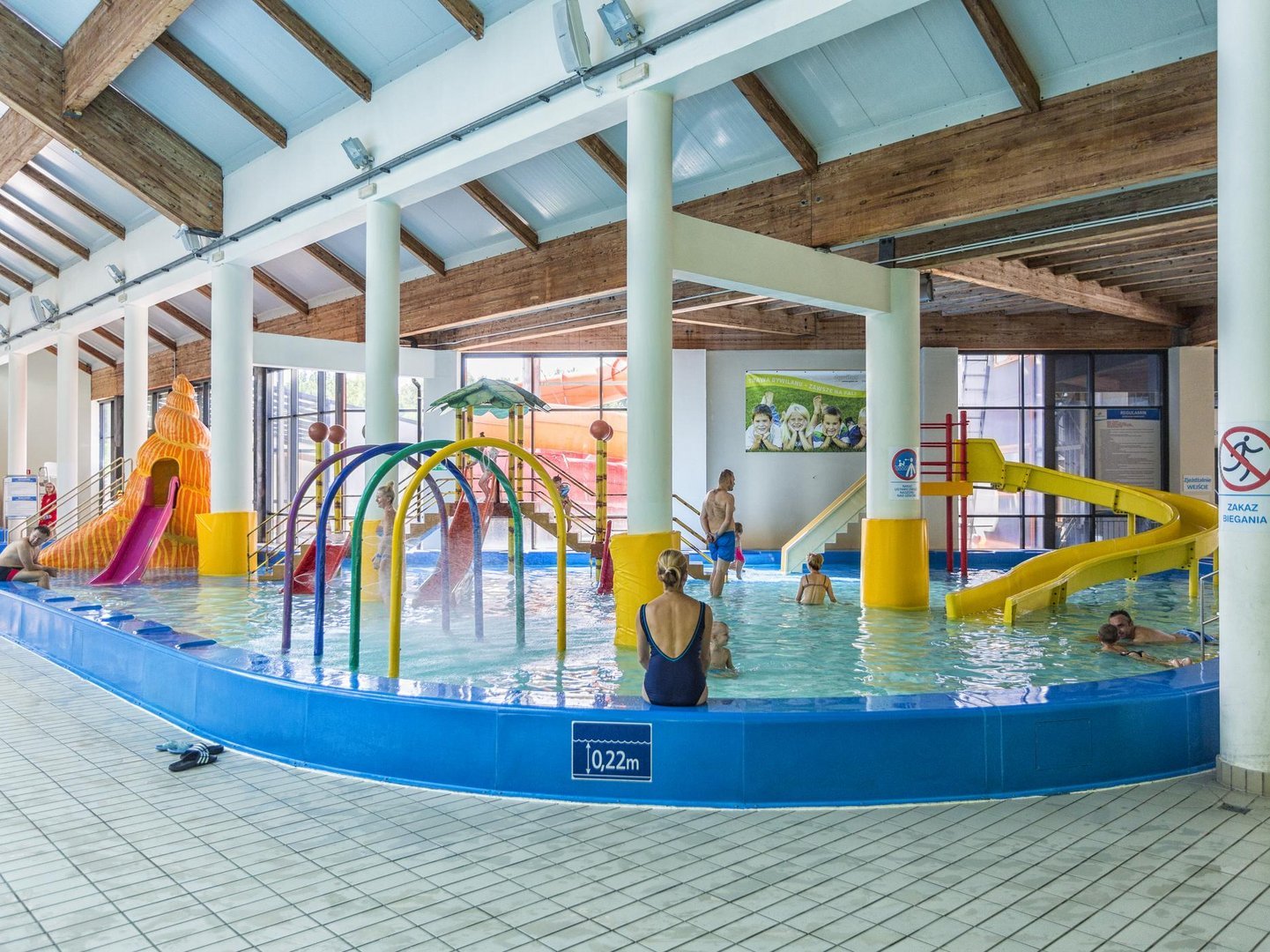 Panorama wodnego placu zabaw z muszlą. Na pierwszym planie mama nadzorująca zabawę dzieci, kolorowe kurtyny wodne, w tle dom na palach. Po prawo żółta zjeżdżalnia, a po lewo zjeżdżalnia-muszla.