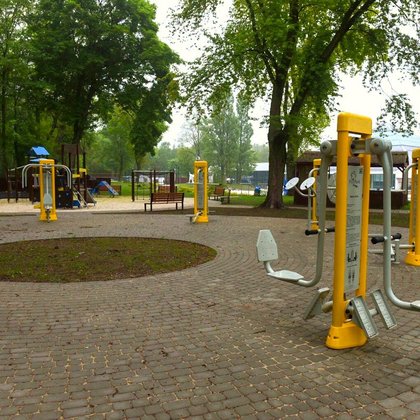 Siłownia zewnętrzna usytuowana w części parkowego Aquaparku FALA. Na szarej kostce brukowej stoją żółto-szare sprzęty. W tle plac zabaw dla dzieci i jedna z hal-piramid Aquaparku.