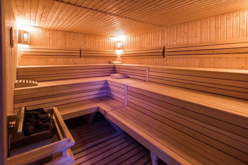Wnętrze wewnętrznej sauny fińskiej, po lewej stronie przy drzwiach piec z ciemnymi kamieniami, naprzeciw i po prawej stronie trzy rzędy siedzisk. Cała sauna zabudowana drewnem.
